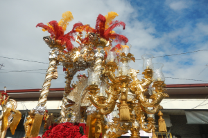 Ein großer goldener und roter Rosenmontagszug, geschmückt mit Blumen und Verzierungen, mit einem Gebäude und Strommasten mit Drähten im Hintergrund unter einem bewölkten Himmel.