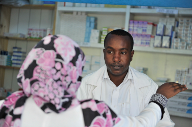 Eine Apothekerin in einem weißen Kittel steht vor einer sitzenden Frau in einer Apotheke, mit Regalen voller Schachteln und Gegenständen im Hintergrund.