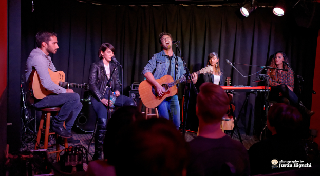 Fünf Personen auf der Bühne, zwei spielen Gitarren, alle mit Mikrofonen und Ständern, mit einem Vorhang und Lichtern im Hintergrund.