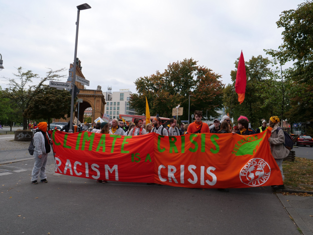 Eine Gruppe von Menschen marschiert eine von Bäumen gesäumte Straße entlang, die ein Banner mit der Aufschrift "Klima-Krise ist eine Krise" trägt, mit Fahrzeugen und Gebäuden im Hintergrund unter einem klaren blauen Himmel.