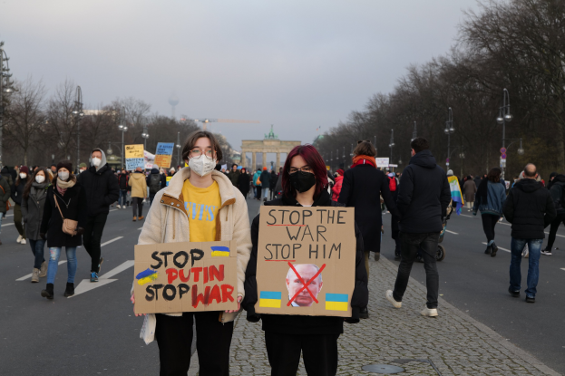 Eine Gruppe von Menschen, die auf der Straße in Berlin, Deutschland, entlanggehen, Masken tragen und Schilder halten, auf denen "Stoppt den Krieg" steht, mit Bäumen und Laternen an der Straße, einem Bogen im Hintergrund und einem klaren blauen Himmel.