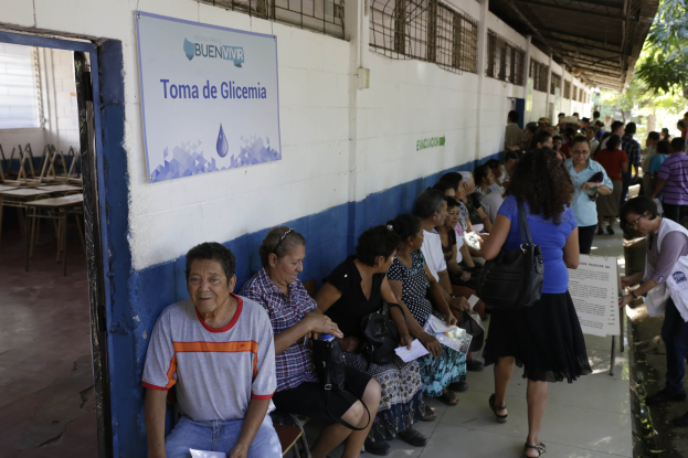 Menschen sitzen auf Bänken vor einem Gebäude mit Papieren und Taschen, Warteschlange an einer Urne mit einer Tafel an der Wand, Tische, Stühle, Bäume und ein Schuppen im Hintergrund.