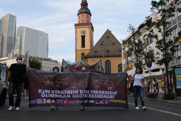 Eine Gruppe maskierter Personen marschiert auf einer Straße und hält ein Banner hoch, mit einem geparkten Auto auf der linken Seite, Gebäuden, Bäumen, Schildern, Polen und einem Kirchturm im Hintergrund unter einem klaren blauen Himmel.