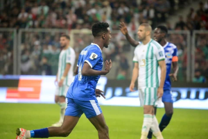 Ein Fußballspieler in einem blauen und weißen Trikot feiert ein Tor, umgeben von einer Gruppe in weißen und grünen Trikots, mit einer weißen und blauen Tafel und einem Geländer im unscharfen Hintergrund.