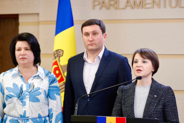 Zwei Frauen und ein Mann stehen an einem Podium vor einer ukrainischen Flagge, wobei der Mann einen schwarzen Blazer und ein weißes Hemd trägt und die Frau rechts ein aschfarbenes Kleid trägt und über die Ukraine-Krise diskutieren.