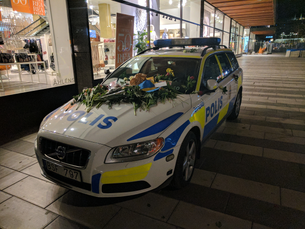 Ein mit Blumen und Blättern geschmückter Polizeiwagen steht nachts vor einem Geschäft mit Glaswänden, mit Schaufensterpuppen in Kleidern, Texttafeln und anderen Gegenständen im Inneren und Bäumen und Gebäuden im Hintergrund.