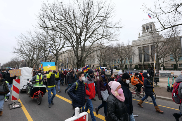 Eine große Gruppe von Menschen marschiert auf einer Stadtstraße in Washington, D.C. mit Schildern und Transparenten, einige fahren Fahrräder, unter einem klaren blauen Himmel.