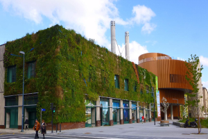 Ein großes Gebäude mit einer grünen Wand an der Seite, umgeben von Straßeninfrastruktur, Fahrzeugen, Fußgängern, Bäumen und einem bewölkten Himmel, Teil des Green Building Projects.