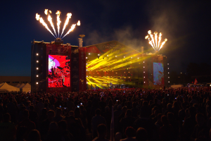 Eine große Menschenmenge auf einem Musikfestival schaut auf eine hell erleuchtete Bühne, mit Zelten links und Feuerwerk am dunklen Himmel dahinter.