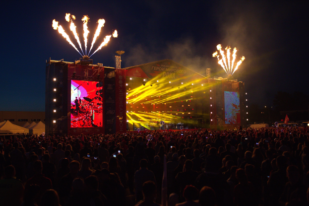 Eine große Menschenmenge auf einem Musikfestival schaut auf eine hell erleuchtete Bühne, mit Zelten links und Feuerwerk am dunklen Himmel dahinter.