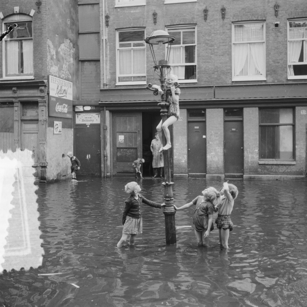 Kinder spielen in einer überfluteten Straße in Amsterdam, mit einem Pfahl und einer Lampe in der Mitte und Gebäuden mit Fenstern, Türen und Texttafeln im Hintergrund.