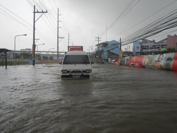 Ein Van fährt durch eine überflutete Stadtstraße mit Strommasten, Laternenmasten und Gebäuden auf beiden Seiten, eine Brücke im Hintergrund und der Himmel darüber.