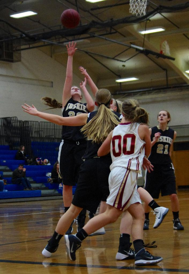 Mehrere Frauen in Trikots auf einem Basketballfeld, mit einem Basketball und Netz sowie Zuschauern im Hintergrund unter Deckenbeleuchtung.
