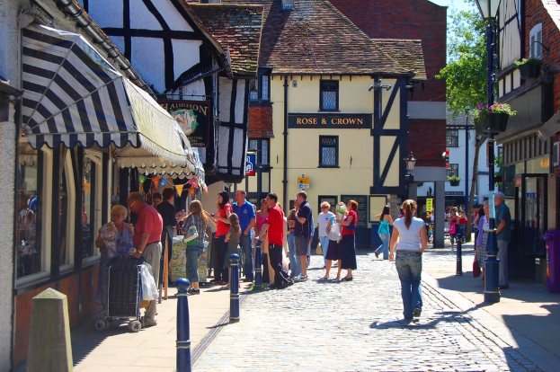 Eine Straße mit Pfosten und Gebäuden auf beiden Seiten, Menschen auf der linken Seite mit einem Karren, ein Mann und eine Frau auf der rechten Seite in der Nähe eines lila Müllcontainers, mit Gebäuden, Bäumen und Blumentöpfen im Hintergrund.