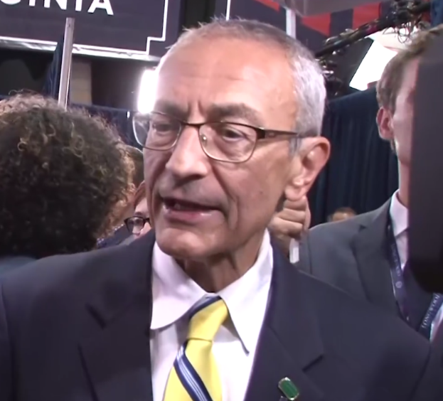 Ein Mann im Anzug und Krawatte mit Brille spricht zu Reportern, umgeben von einer Gruppe von Menschen, mit einem Mikrofonständer vor ihm und einem beleuchteten Hintergrund mit einer Tafel, auf der Text steht.