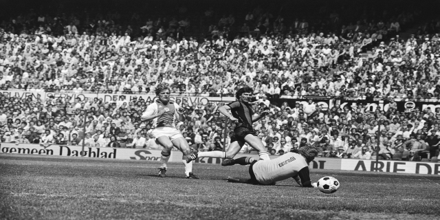 Schwarzes Foto von Männern, die auf einem Feld Fußball spielen, mit Zuschauern im Hintergrund und einer Tafel mit der Aufschrift "Bundesliga 1978-79 Ajax 1:1 Hannover 96."