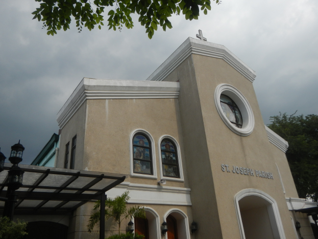St. Joseph Parish Church in Manila, Philippines, a building with glass windows and doors, surrounded by trees and plants, with a light pole on the left side and a cloudy sky in the background.