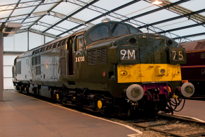 Ein Zug befindet sich auf den Schienen neben einer Plattform mit einem an der Decke befestigten Pfosten, der Metallstangen mit Lampen trägt.