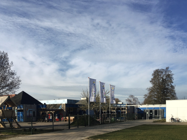Großes Gebäude mit Metallzaun, Rasenfläche, Weg, Bäumen, Pfosten, Bannern, Fahrrad, nahen Häusern und bewölktem Himmel, identifiziert als neuer Spielbereich für Kinder an der Schule.