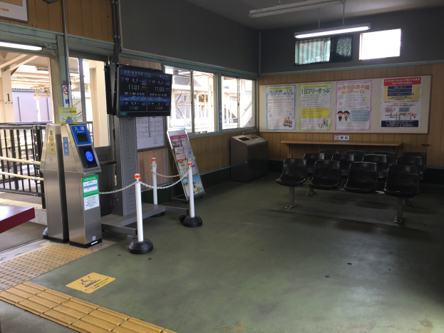 Ein belebter Bahnhofseingang mit Sitzgelegenheiten, einem Service-Schalter, Ticketautomaten, Kettenzäunen, Informationsanzeigen, Wandplakaten, Deckenleuchten und Geländern.