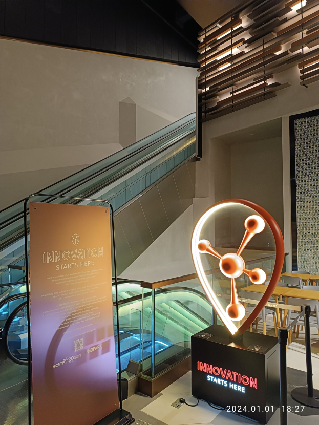 Großes Schild mit der Aufschrift "Innovation beginnt hier" vor einem Gebäude mit einer Treppe im Hintergrund, Tischen und Stühlen auf der rechten Seite und einer Tafel auf der linken Seite, die die Teilnahme an der HSTP20 Expo anzeigt.