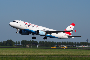 Ein Airbus A320-200 der Austrian Airlines startet vom Frankfurter Flughafen, mit grünem Gras, Bäumen, Gebäuden und einem Turm im Hintergrund, unter einem klaren Himmel.