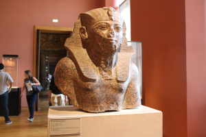 Ein Büste eines ägyptischen Pharaos ist in einem Museum ausgestellt mit einer Texttafel, einer Gruppe von Menschen, einer Frau mit einer Tasche, einer Tür, einer Wand und einer Decke mit Lampen.