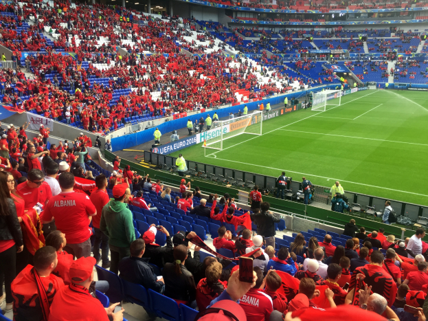 Ein volles Stadion mit sitzenden und stehenden Zuschauern, Bannern, einem Torpfosten und Deckenleuchten, die Menge erwartet gespannt den Beginn eines Füßballspiels.