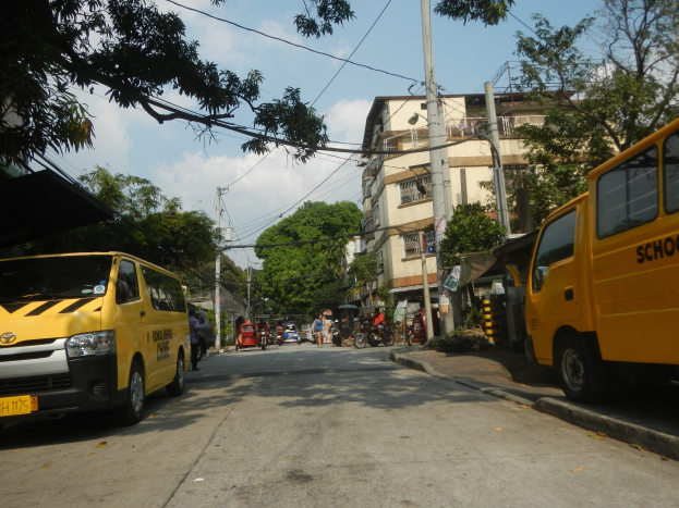 Ein gelber Schulbus fährt eine Straße entlang, die von hohen Gebäuden, Bäumen und Strommasten mit Drähten gesäumt ist, mit Menschen auf Fahrrädern und anderen Fahrzeugen auf der Straße, unter einem bewölkten Himmel.