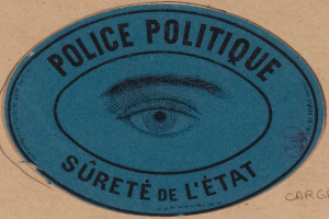 Eine braune Pappkiste mit einem blauen Aufkleber, auf dem "Polizei Politique Sûreté de l'État" und ein Auge in der Mitte steht.