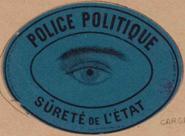 Eine braune Pappkiste mit einem blauen Aufkleber, auf dem "Polizei Politique Sûreté de l'État" und ein Auge in der Mitte steht.