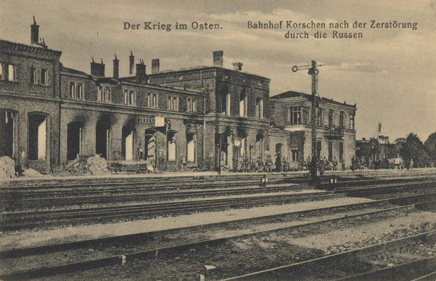 Schwarzes und weißes Foto der Bahnstation Korschen in Ostern, Deutschland, mit Bahnschienen, Bahnhofsgebäuden, Menschen, Bäumen und einem Himmel-Hintergrund mit Text oben und unten.