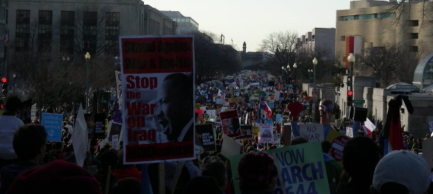 Eine Gruppe von Menschen demonstriert auf einer Straße und hält Plakate, mit Gebäuden, Bäumen und Laternen im Hintergrund bei klarem Himmel.