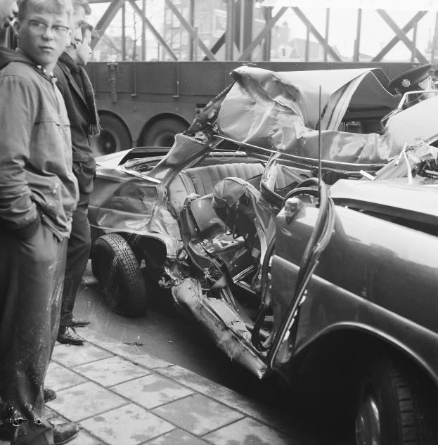 Schwarz-weißes Bild eines verunglückten Autos am Straßenrand mit mehreren Menschen in der Nähe, einem Lkw und einer Brücke im Hintergrund.