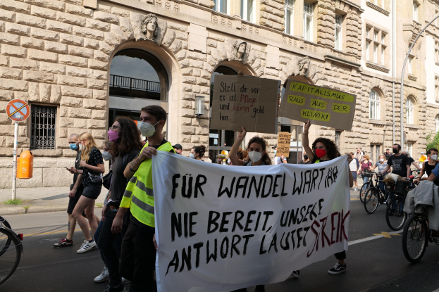 Eine Gruppe von Menschen marschiert auf einer Straße, einige halten Schilder und andere fahren Fahrräder, im Hintergrund ein Gebäude mit Fenstern, Bögen, Säulen und Skulpturen.