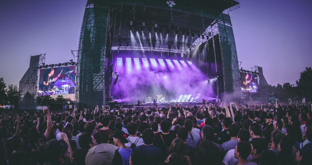 Eine große Menschenmenge bei einem Musikfestival mit Künstlern auf einer beleuchteten Bühne, umgeben von Bildschirmen, vor einem Gebäude, Bäumen und einem klaren blauen Himmel.