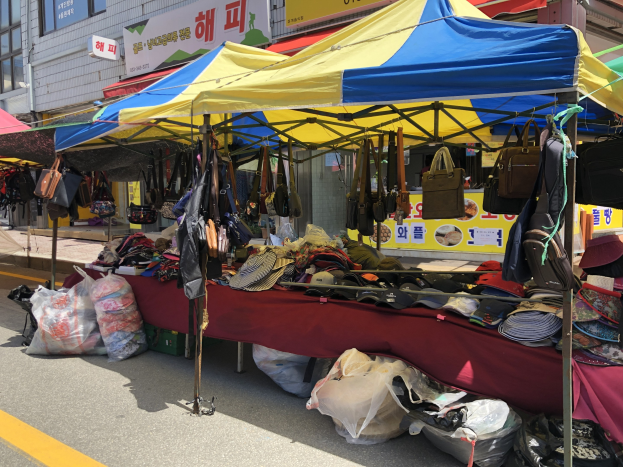 Ein Markt mit Zelten, die verschiedene Gegenstände wie Taschen und Hüte präsentieren, vor einem Hintergrund aus Gebäuden mit Fenstern und Texttafeln.