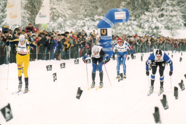 Gruppe von Skifahrern in Helmen, Handschuhen und Skibrillen, die eine schneebedeckte Piste hinabfahren, mit Zuschauern, Tannen und einem Bogen im Hintergrund.