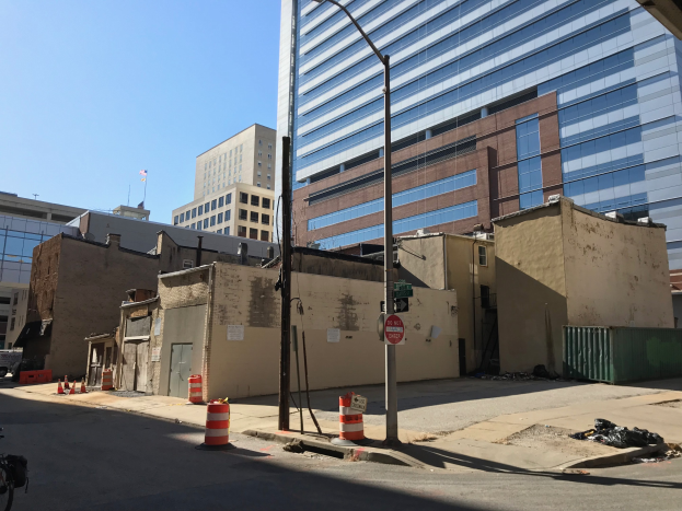 Eine Stadtstraße mit einem geparkten Fahrrad, umgeben von Gebäuden, Straßeninfrastruktur, fahrenden Fahrzeugen und einem demolierten Gebäude im Hintergrund unter einem klaren Himmel.
