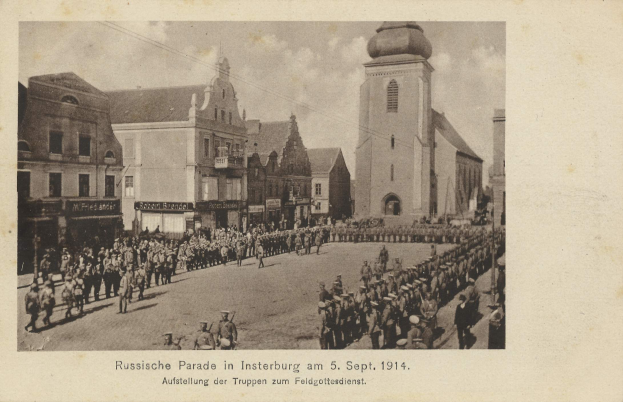 Ein Schwarz-Weiß-Foto eines Umzugs in Insterburg am 5. September 1914 mit vielen Menschen, Gebäuden und bewölktem Himmel sowie einem Text unten.