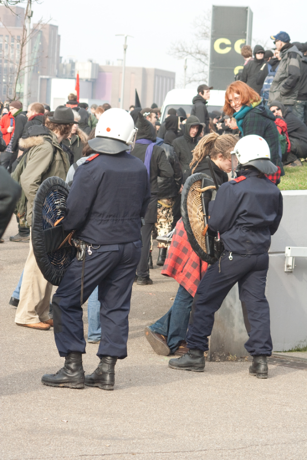 Eine Gruppe von Menschen, die auf einer Straße gehen, mit zwei Personen vorne, die wie Polizei aussehen, Gebäuden im Hintergrund und Boden unten.