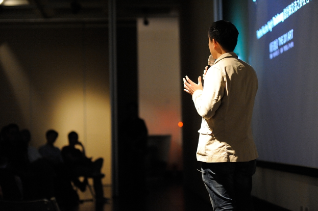 Eine Person steht auf einer Bühne und spricht in ein Mikrofon, mit einem Bildschirm dahinter, ein paar sitzende Personen davor und einer Lampe neben ihnen.