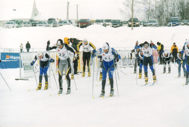 Eine Gruppe von Menschen in Skiausrüstung fährt eine schneebedeckte Piste hinunter, mit Bannern, Menschen, Fahrzeugen, Pfählen, Bäumen und einem klaren blauen Himmel im Hintergrund.