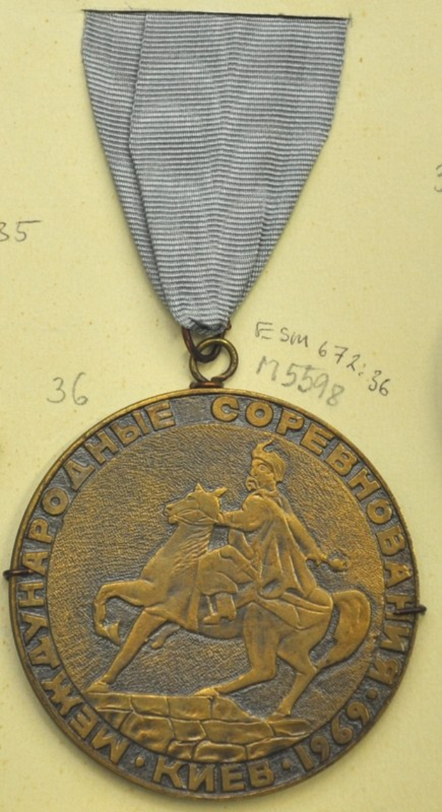 Goldmedaille mit galoppierendem Pferd und Reiter, befestigt an einem Dokument mit Text und Zahlen.