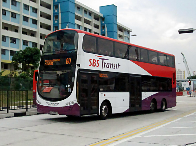Ein Elektrobus ist in der Mitte des Bildes zu sehen, im Hintergrund ein Gebäude, Bäume vor dem Gebäude, ein Zaun auf der rechten Seite des Fußwegs und Himmel oben.