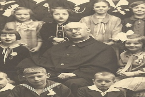 Ein Schwarz-Wei├č-Foto eines strengen Mannes, der in der Mitte sitzt und von einer Gruppe Kinder umgeben ist, die bewundernd zu ihm aufschauen.