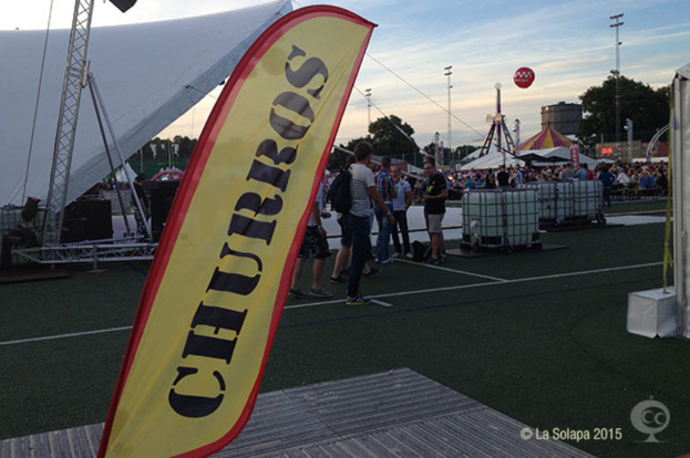 Ein buntes Karnevalszelt mit Zelten, einem zentralen Mast, Menschen, Bäumen, Strommasten, einem Himmel, Straßenbahnen auf der Straße und einem Eisenständer auf der linken Seite.