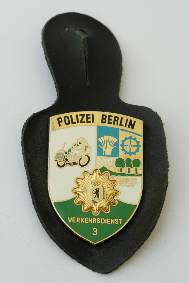 Ein schildförmiges Polizeibadgebnis mit "Polizei Berlin" in schwarzer fettgedruckter Schrift, das ein weißes Stern auf einem blauen Hintergrund und einen weißen Rand hat, auf einer Oberfläche.
