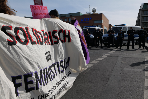Gruppe von Menschen marschiert auf einer Straße mit einem "Solidarität und Feminismus"-Schild, mit geparkten Fahrzeugen, Gebäuden, einer Schüsselantenne und einem klaren blauen Himmel im Hintergrund.