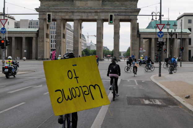 Eine Gruppe von Menschen auf Fahrrädern fährt eine Straße entlang vor dem Reichstagsgebäude in Berlin, Deutschland, trägt Helme und hält ein gelbes Schild, mit Laternenpfählen, Verkehrsampeln, Gebäuden, Bäumen und einem klaren blauen Himmel im Hintergrund.